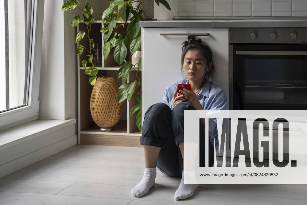 Aufgeregte, düstere junge asiatische Frau, die ein Handy in der Hand hält und traurig auf den Handybildschirm schaut. Besorgtes Mädchen sitzt auf dem Boden in der Küche mit Smartphone in den Händen und wartet auf einen wichtigen Anruf Upset gloomy young Asian woman holding cellphone, looking at mobile phone screen feeling sad. Concerned girl sitting on floor in kitchen with smartphone in hands waiting for important call