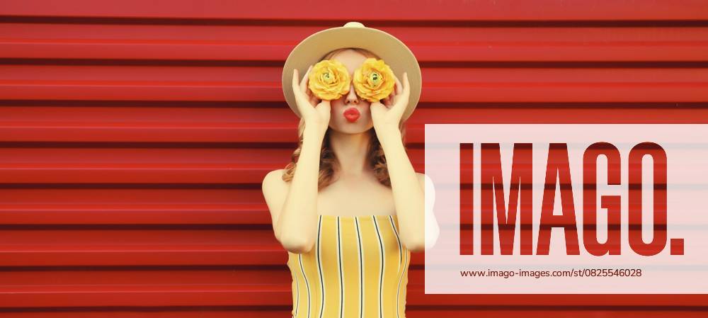Sommer Porträt der stilvollen jungen Frau bläst Kuss mit gelben Blütenknospen auf der Suche nach etwas in runden Strohhut auf rotem Hintergrund Summer portrait of stylish young woman blowing kiss with yellow flower buds looking for something in round straw hat on red background