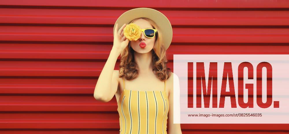 Sommer Porträt der stilvollen jungen Frau bläst Kuss mit gelben Blütenknospen auf der Suche nach etwas in runden Strohhut auf rotem Hintergrund Summer portrait of stylish young woman blowing kiss with yellow flower buds looking for something in round straw hat on red background