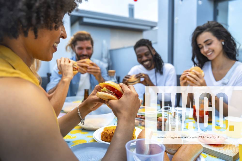 Fröhliche, multiethnische Freunde genießen leckere Burger bei einer lebhaften Grillparty auf dem Dach, bei der viel gelacht wird und man zusammenkommt Cheerful multi ethnic friends savoring delicious burgers while enjoying a lively rooftop barbecue party filled with laughter and togetherness