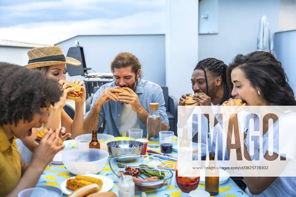 Fröhliche multiethnische Freunde genießen leckere Burger bei einer lebhaften Grillparty auf dem Dach und feiern gemeinsam den Sommer Cheerful multi ethnic friends savoring delicious burgers while enjoying a lively rooftop barbecue party, celebrating summer together