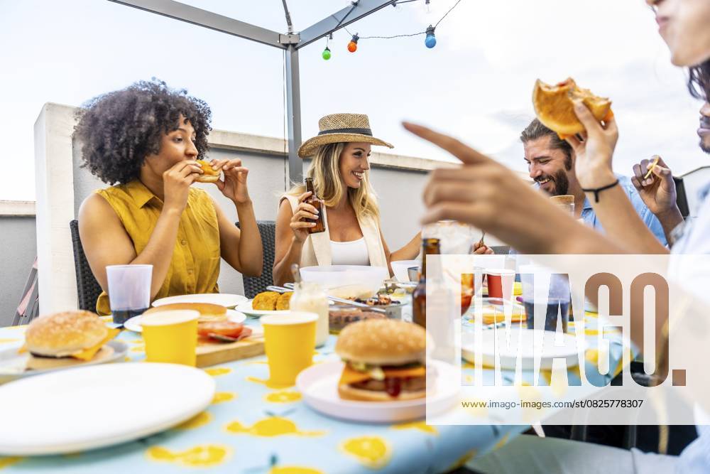 Fröhliche Freunde genießen Burger und Bier und feiern ihre Zweisamkeit auf einer Dachterrasse mit Lichterketten Happy friends enjoying burgers and beer, celebrating togetherness on a rooftop with string lights