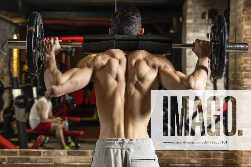 Rückenansicht eines jungen Bodybuilders, der seine Muskeln mit einer Langhantel vor einem Spiegel im Fitnessstudio anspannt Back view of young bodybuilder flexing muscles with barbell in front of mirror at gym