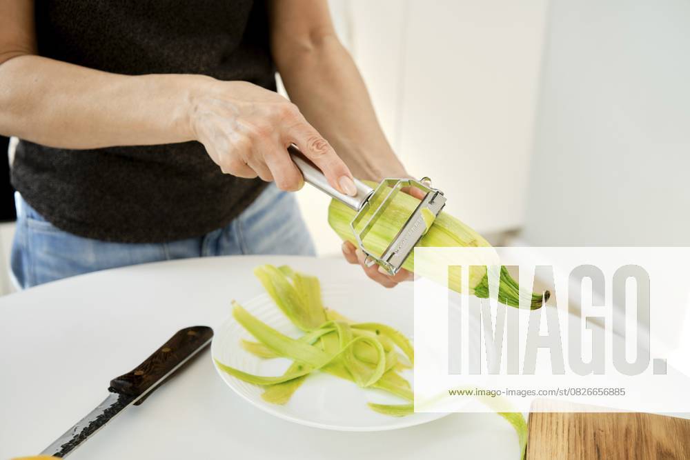 Eine Person schält eine Zucchini in einer hellen Küche. Frische Zucchini werden in einer Hand gehalten, während ein Schäler die Schale entfernt A person is peeling a zucchini in a bright kitchen. Fresh zucchini is held in one hand while a peeler removes the skin