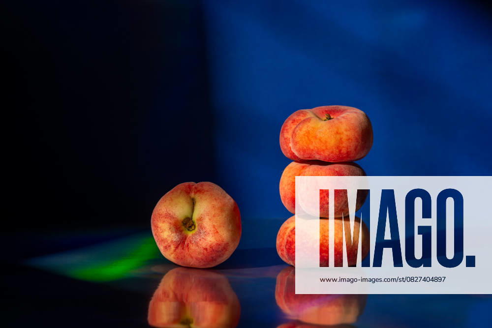 A stack of three flat peaches and one beside it sit on a reflective surface. The dark blue background enhances their vibrant colors and textures, creating a striking composition. Model Released Ellsy_ID30337_1023860_001
