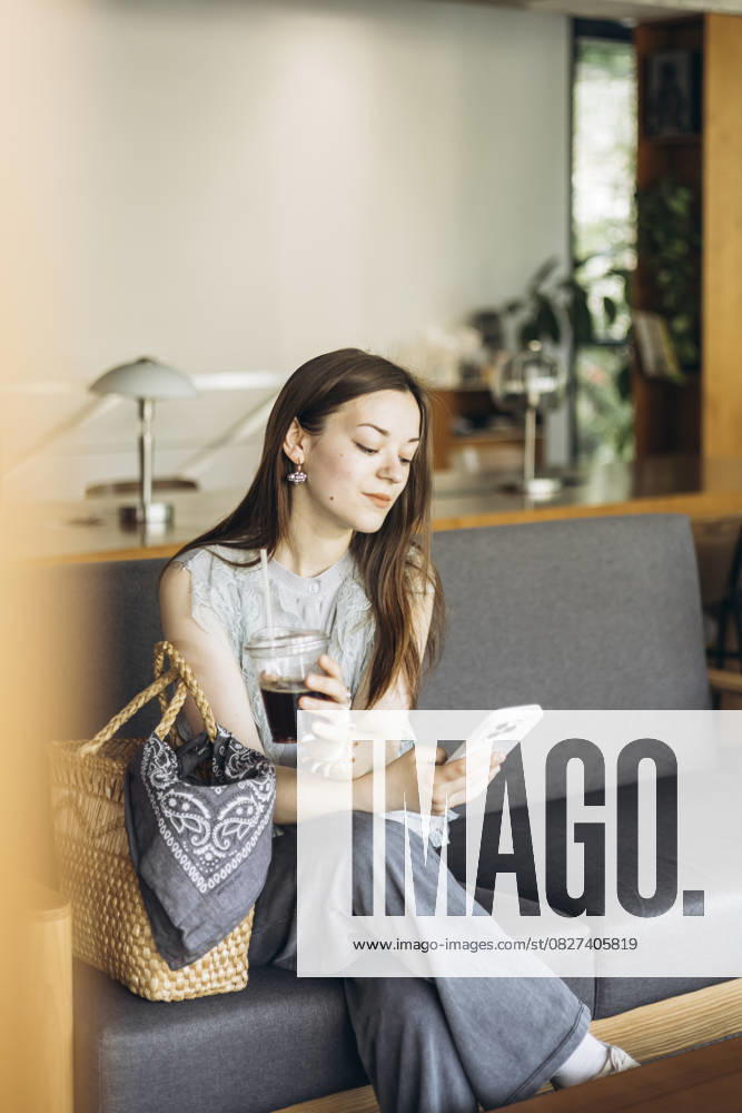 IMAGO Creative, Food, Trend, Iced Coffee A woman sits comfortably on a sofa in a cozy cafe, holding an iced coffee in one hand and a smartphone in the other, epitomizing relaxation and modern lifestyle. Model Released HannaYakimenko_ID30226_1019070_009