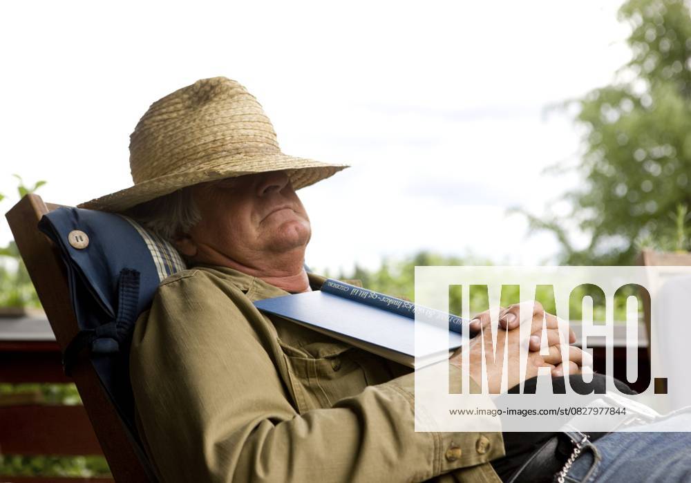 Älterer Mann ruht sich auf einer Veranda aus. Gesicht durch Strohhut verdeckt. Hält ein Buch Elderly man resting on veranda. Face covered by straw hat. Holding book
