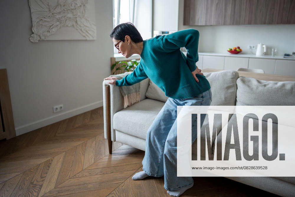 Ältere Frau, die sich wegen plötzlicher Wirbelsäulenschmerzen schmerzhaft an den Rücken klammert. Bandscheibenvorfall, Gelenkerkrankung Senior woman gripping lower back in severe pain from sudden spinal attack. Slipped disc, arthritic condition, muscle spasm, acute discomfort, standing difficulty, indoor scene, health emergency (License=RF) ,model released, Symbolfoto