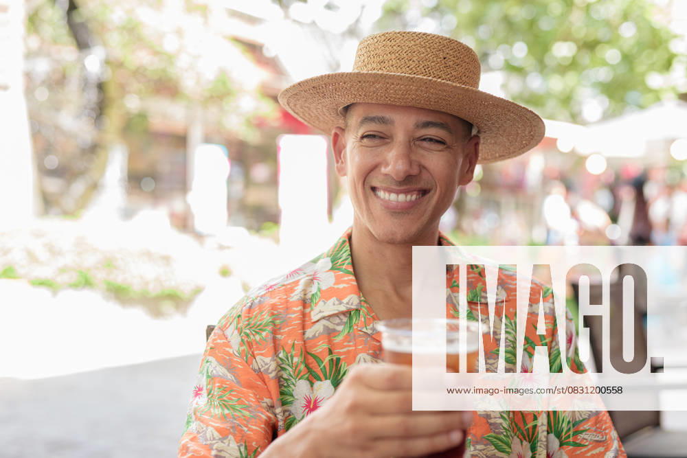 IMAGO Creative, Food, Trend, Iced Coffee Hispanischer Mann mit Eiskaffee in einem Straßencafé, der einen Panamahut und ein kurzärmeliges Hemd trägt A Hispanic male tourist enjoying an iced coffee while sitting at an outdoor cafe on a sunny day, wearing a Panama hat and short sleeve shirt. Casual summer beverage scene. (License=RF) ,model released, Symbolfoto