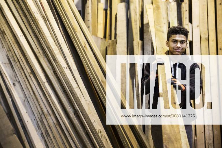 IMAGO Creative: Nächste Generation, Sozial- und klimabewusst Young man wearing work gloves standing next to a stack of wooden planks in a warehouse, carrying long pieces of wood, looking at camera. Young man wearing work gloves standing next to a stack of wooden planks in a warehouse, carrying long pieces of wood, looking at camera. 40694