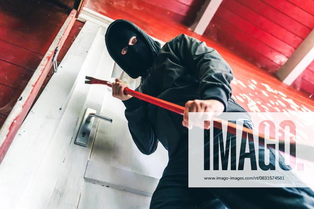 Masked burglar stands in front of a front door with a crowbar. Symbolic image for burglary and robbery