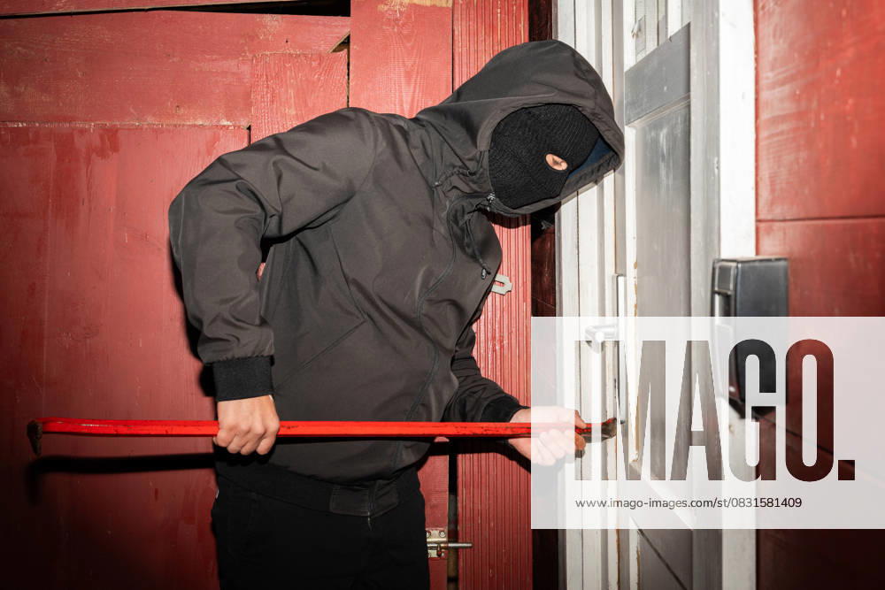 A masked man tries to break down a front door with a crowbar. Symbolic image of the danger of burglaries and the risk to houses and apartments from crime