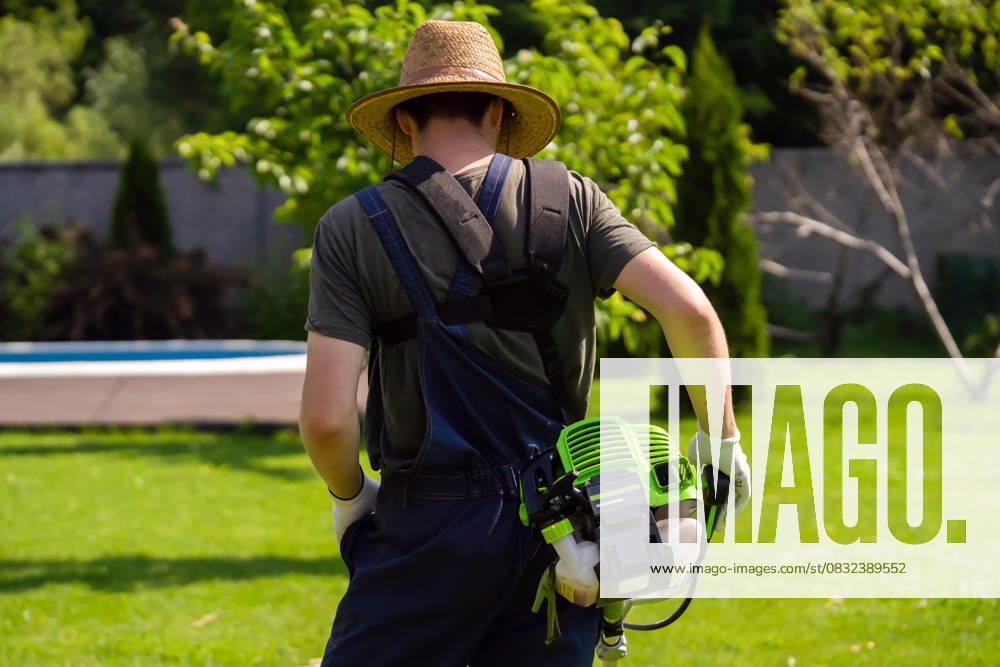 A man with a lawnmower cares for the grass in the backyard. A young man in a straw hat is mowing a lawn with a lawn mower in his beautiful green floral summer garden. A professional gardener with a lawnmower cares for the grass in the backyard., model released, , 65024838.jpg RECORD DATE NOT STATED