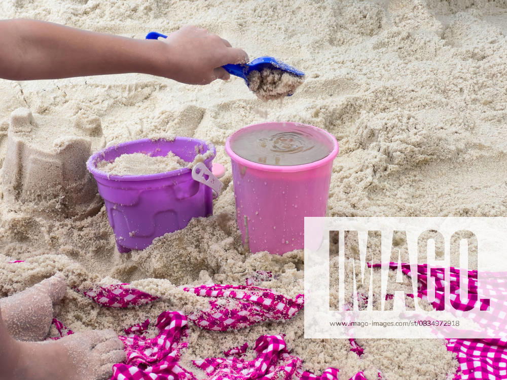 Child Building Sandcastles with Colorful Buckets at the Beach layful scene of a child s hand scooping wet sand into a bright pink and purple bucket using a blue plastic shovel. The sandy beach is scattered with footprints, sandcastles, and a red and white checkered fabric, enhancing the summer vacation vibe. This image captures childhood joy, outdoor fun, and beachside adventures, making it perfect for travel and lifestyle content., 95429814.jpg RECORD DATE NOT STATED