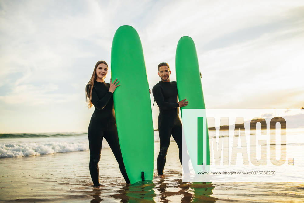 Couple Standing With Surfboards in Shallow Ocean Water at Sunset A couple in black wetsuits stands together in the shallow ocean water with their green surfboards. They are smiling and looking directly at the camera. The sun is setting behind them, model released, , 60769338.jpg RECORD DATE NOT STATED