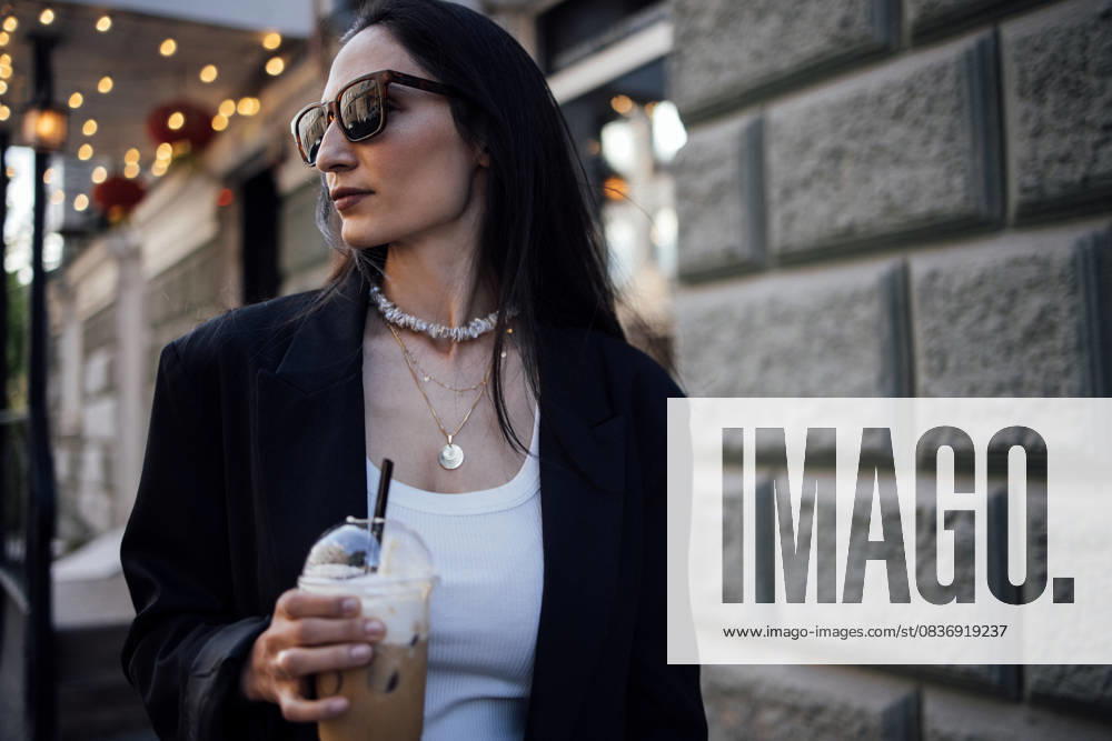 IMAGO Creative, Food, Trend, Iced Coffee Stilvolle Frau mit langen dunklen Haaren, die eine Sonnenbrille und einen schwarzen Blazer trägt und einen Eiskaffee in der Hand hält, geht in einer trendigen städtischen Umgebung spazieren und präsentiert moderne Mode und Lebensstil Stilvolle Frau mit langen dunklen Haaren, die eine Sonnenbrille und einen schwarzen Blazer trägt und einen Eiskaffee in der Hand hält, geht in einer trendigen städtischen Umgebung spazieren und präsentiert moderne Mode und Lebensstil (License=RF) ,model released, Symbolfoto