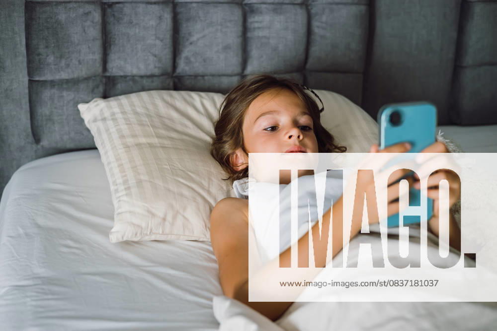 Ein junges Mädchen konzentriert sich auf ihr Smartphone, während es sich im Bett entspannt. Konzept: Bildschirmzeit, digitale Gewohnheiten und modernes Leben zu Hause A young girl focused on her smartphone while relaxing in bed. This photo captures a common scene of childhood and technology, suitable for topics of screen time, digital habits, and modern home life (License=RF) ,model released, Symbolfoto