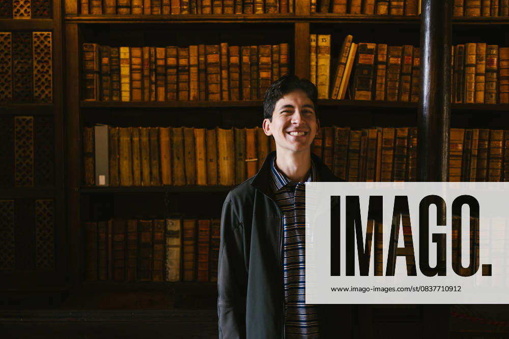 Young adult man smiling in front of book shelf in library Oxford, England, United Kingdom CR_WVNP251008B-1799229-01 ,model released, Symbolfoto