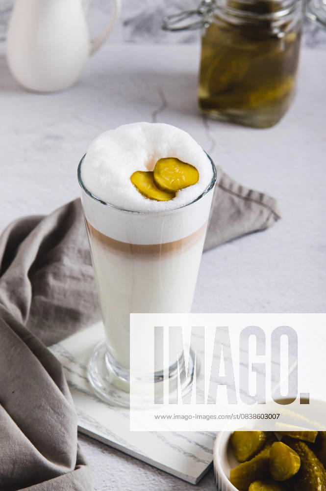 IMAGO Creative, Food, Trend, Iced Coffee Picklatte mit Schaum in einem Glas und Essiggurken in einer Schale auf dem Tisch Vertikalansicht Picklatte mit Schaum in einem Glas und Essiggurken in einer Schale auf dem Tisch Vertikalansicht (License=RF)