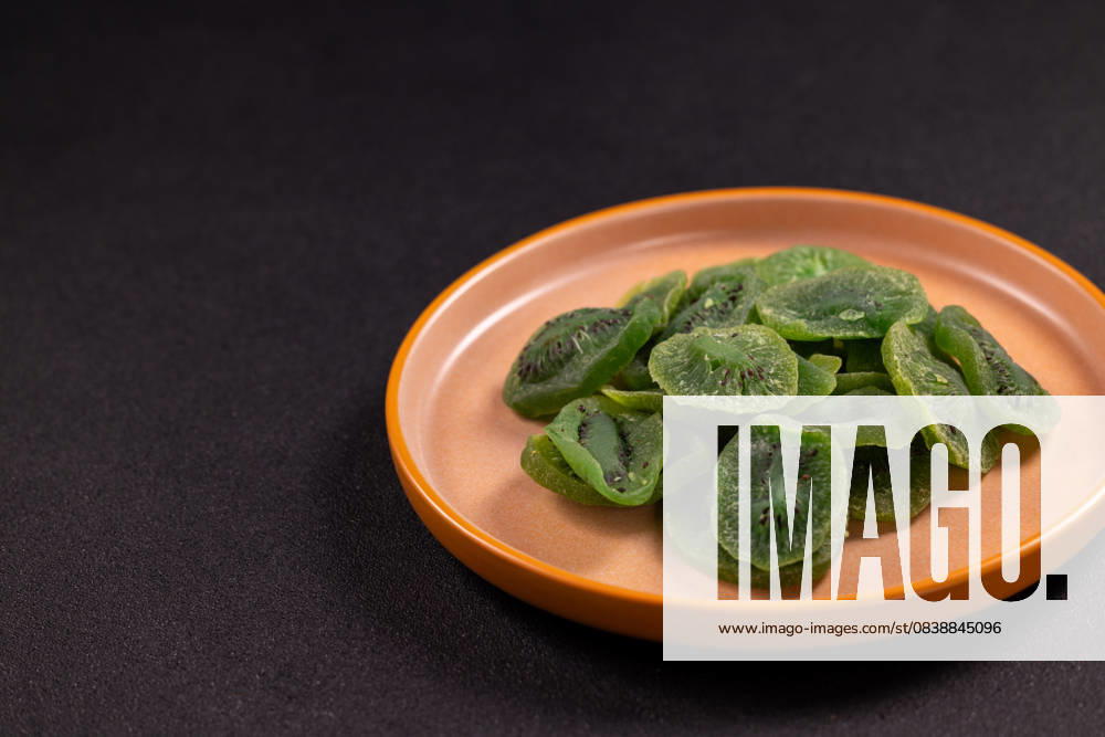 Getrocknete Kiwi-Scheiben auf einem braunen Teller Dried kiwi slices fanned on a rustic brown ceramic plate against a dark backdrop, vibrant green dehydrated fruit showcasing a healthy, sweet, vegan snack or baking ingredient (License=RF)