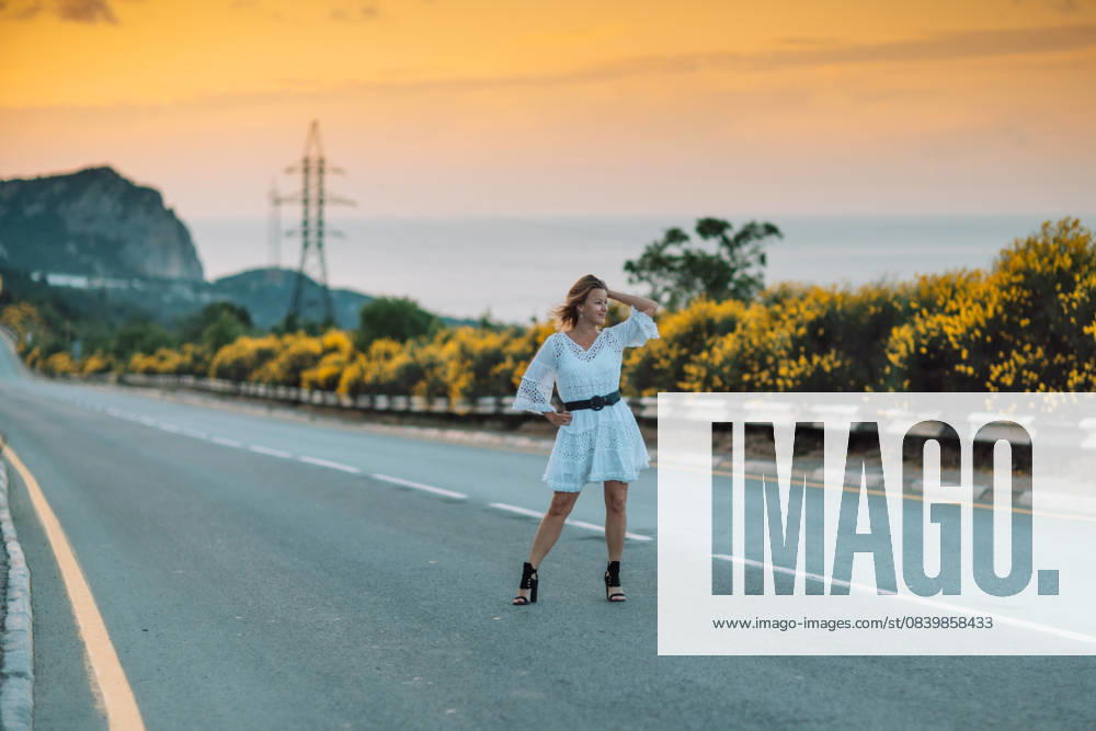 A woman in a white dress is standing on a road, looking at the camera A woman in a white dress is standing on a road, looking at the camera. The scene is set against a backdrop of a beautiful sunset, with the sky turning orange and pink. The woman is enjoying the moment, model released, , 62026546.jpg RECORD DATE NOT STATED