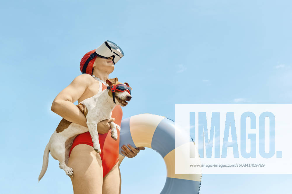 Woman in red swimsuit with virtual reality glasses holding a Jack Russell Terrier and a swimming ring outdoors model released, Symbolfoto property released, CATF00646