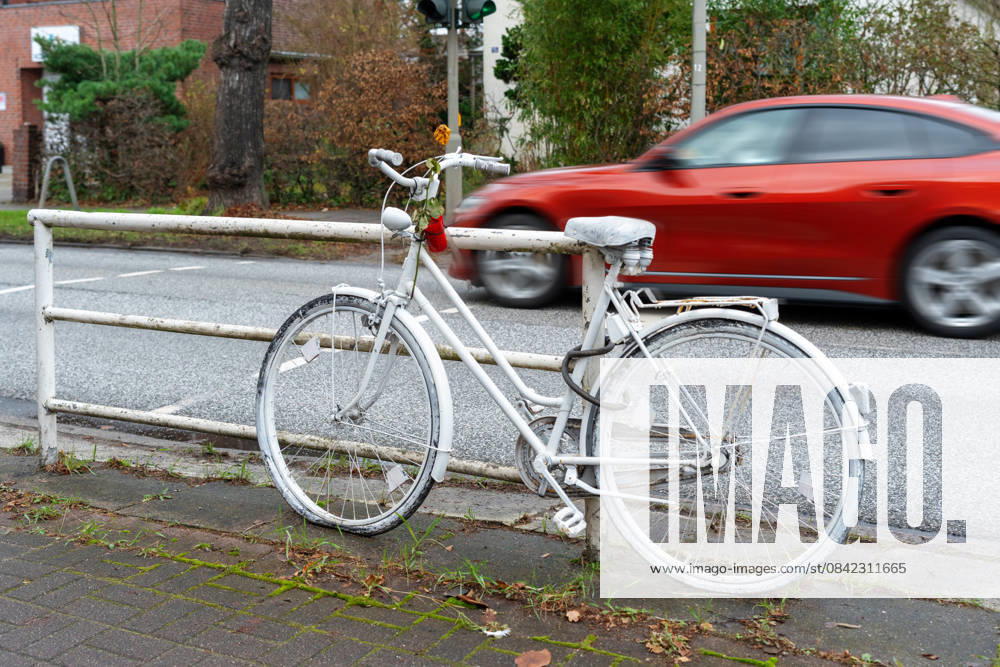 A white ghost bike on the side of the road commemorates a fatal traffic accident. Cars speeding by highlight the dangerous situation cyclists often face in heavy city traffic.