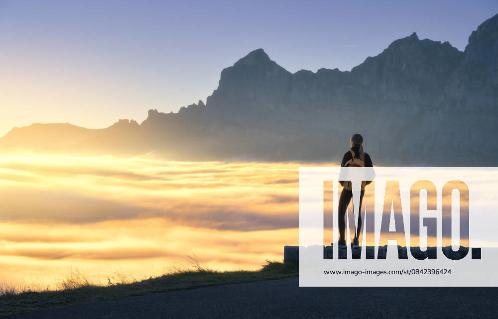 Woman with yellow backpack stands on a viewpoint above a sea of clouds at sunset in Swiss Alps. Girl, sun rays illuminate the alpine mountain peaks and misty valley. Travel in Switzerland. Hiker Model Released DenisBilytskyi_ID32879_1119720_017