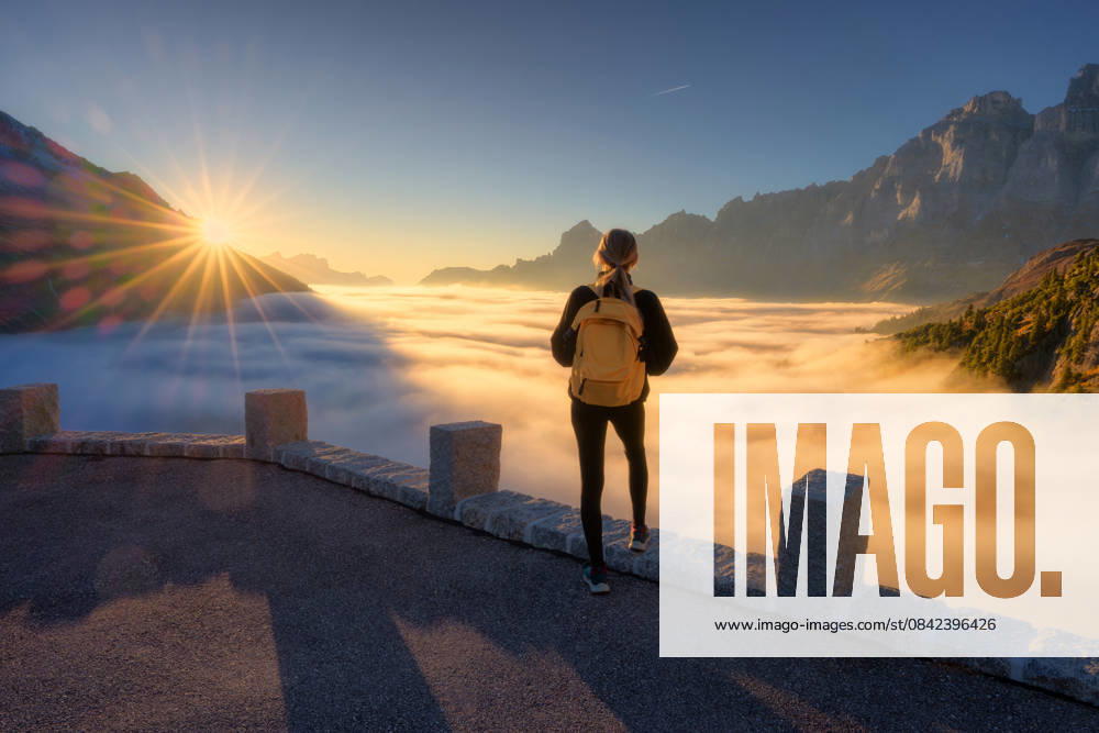 Woman with yellow backpack stands on a viewpoint above a sea of clouds at sunset in Swiss Alps. Girl, sun rays illuminate the alpine mountain peaks and misty valley. Travel in Switzerland. Hiker Model Released DenisBilytskyi_ID32879_1119722_019