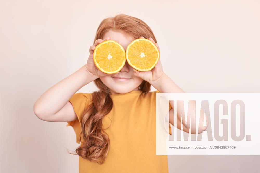 Kaukasisches Kind mit roten Haaren, die die Augen mit orangefarbenen Scheiben bedecken, in verspielter Pose auf beigem Hintergrund Caucasian child with red hair covering eyes with orange slices in playful pose on beige background. (License=RF) ,model released, Symbolfoto