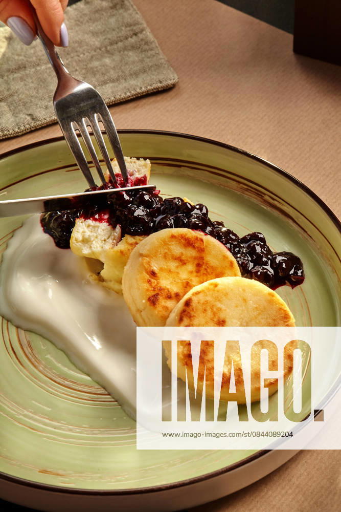 IMAGO Creative, Food, High Protein Frühstück Woman eating syrniki with blueberry jam and sour cream using fork and knife Close-up of delicious syrniki served with blueberry jam and sour cream, being enjoyed by a woman, 97421098.jpg RECORD DATE NOT STATED
