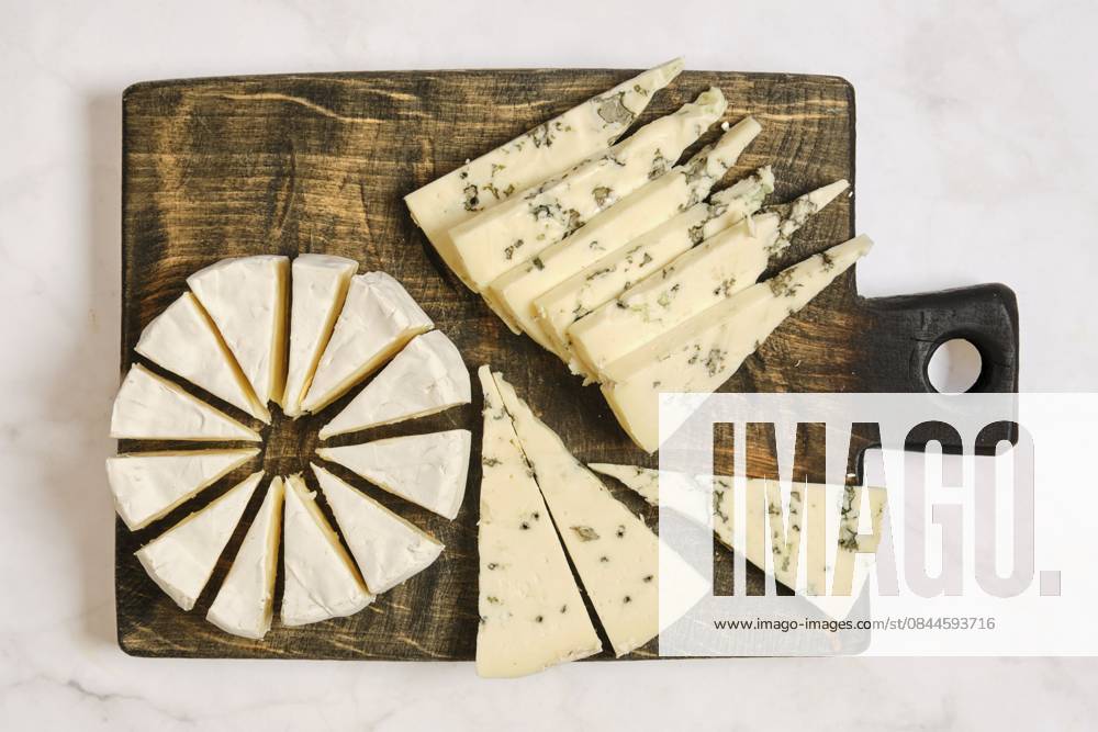 Verschiedene Käsesorten sind auf einem Holzbrett angeordnet. Ein Käse ist rund und in Spalten geschnitten. Ein anderer Typ wird in Dreiecke geschnitten. Die Platte zeigt eine Mischung aus Texturen und Formen Different types of cheese are arranged on a wooden board. One cheese is round and cut into wedges. Another type is sliced into triangles. The platter displays a mix of textures and shapes