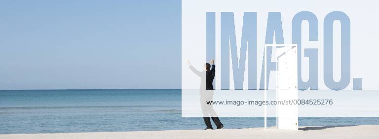 Businessman standing on beach looking at ocean with arms raised, rear view