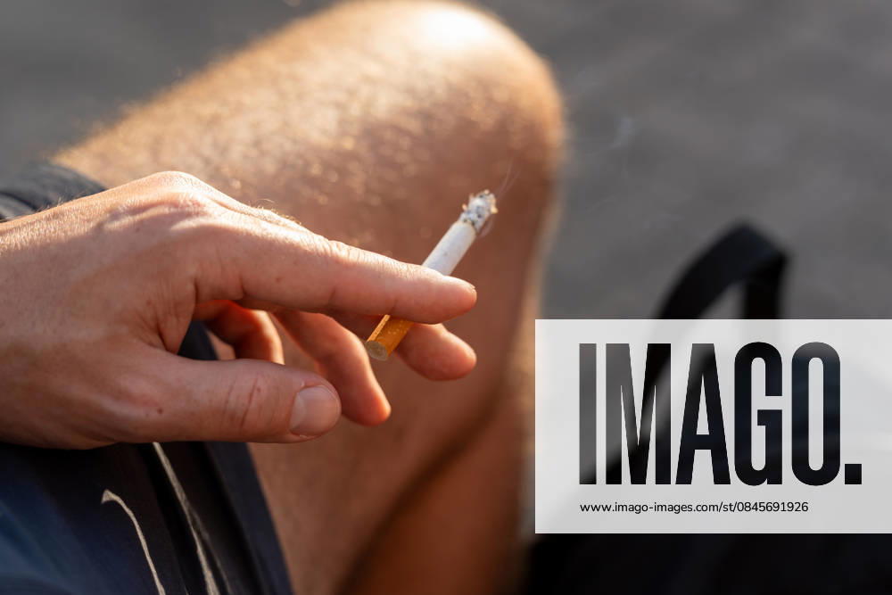 A man smokes a cigarette in swimming trunks outdoors. Smoking is considered a harmful stimulant and poses risks due to nicotine consumption and tobacco smoke. Symbolic image for smoking and addiction