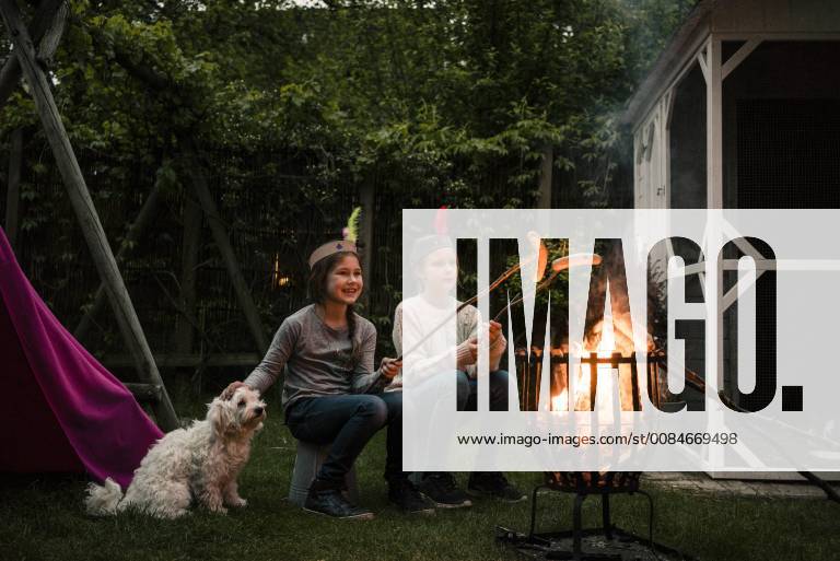 Two girls wearing feather headdress, grilling sausage over camp fire, dog sitting on meadow model released Symbolfoto property released MOEF01216