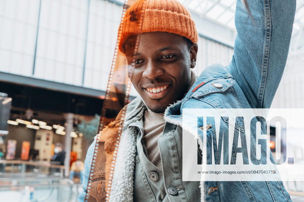 Junge Fotografin untersucht Filmstreifen, lächelnd in einem Einkaufszentrum, trägt orangefarbene Mütze, Jeansjacke, mehrschichtige Freizeitkleidung Young photographer looking at a film strip and smiling while standing in a shopping mall, wearing an orange beanie, denim jacket and layered clothing (License=RF) ,model released, Symbolfoto