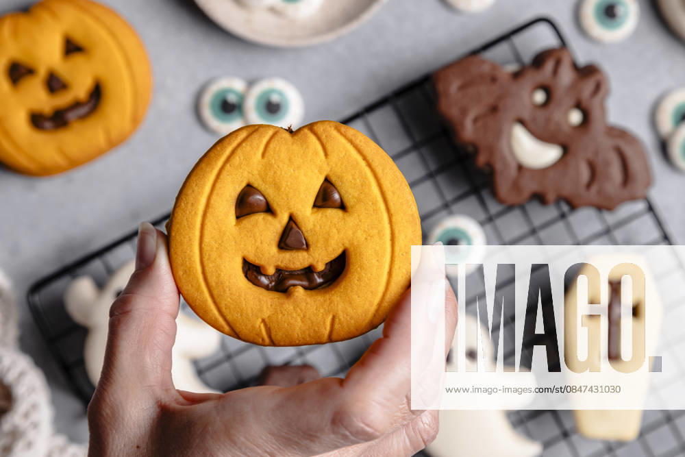Halloween cookies with pumpkin and bat designs Top view of a cropped unrecognizable person holding a pumpkin-shaped Halloween cookie. Background features bat and ghost cookies on a cooling rack over a gray surface. Model Released M-aCreativeStudio_ID32786_1116120_010