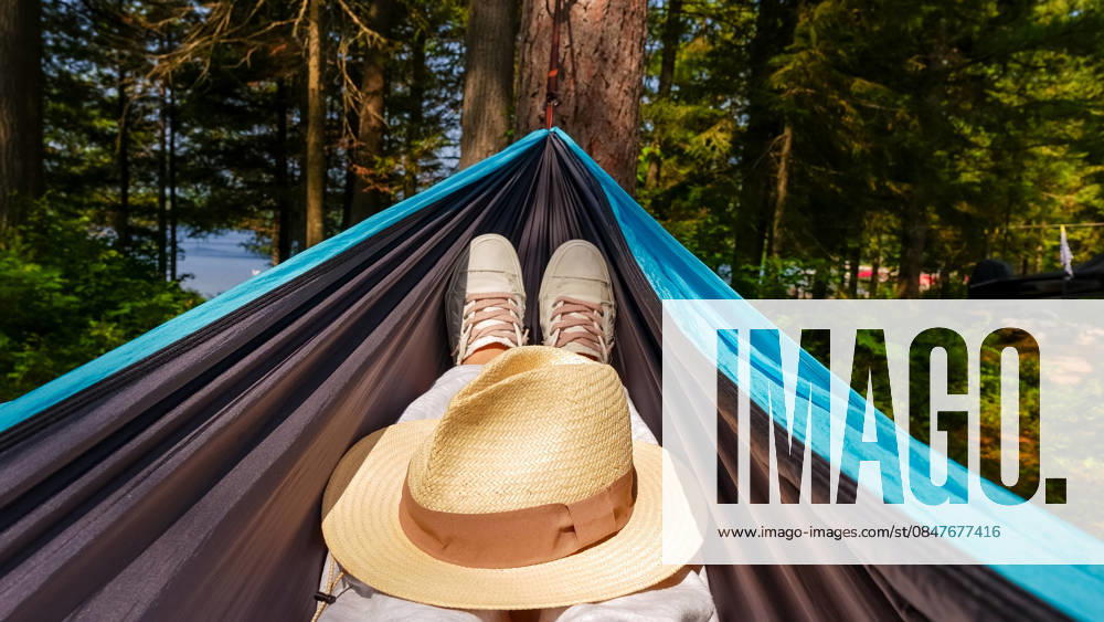 Person, die sich in einer Hängematte entspannt, Turnschuhe und einen Strohhut trägt und von üppig grünen Bäumen in der Nähe eines ruhigen Sees umgeben ist. Individual resting in a hammock with sneakers and a straw hat, nestled among vibrant green trees beside a calm lake (License=RF)