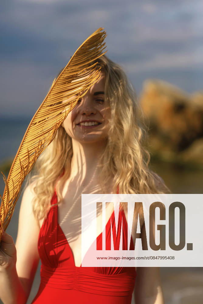Schöne junge Frau mit blonden Haaren posiert am Strand in einem roten Bikini Beautiful young woman with blond hair posing on the beach in a red bikini. She enjoys the wind, smiles. In her hands is a palm leaf, a figured shadow on her face on a summer day. High quality photo. (License=RF) ,model released, Symbolfoto