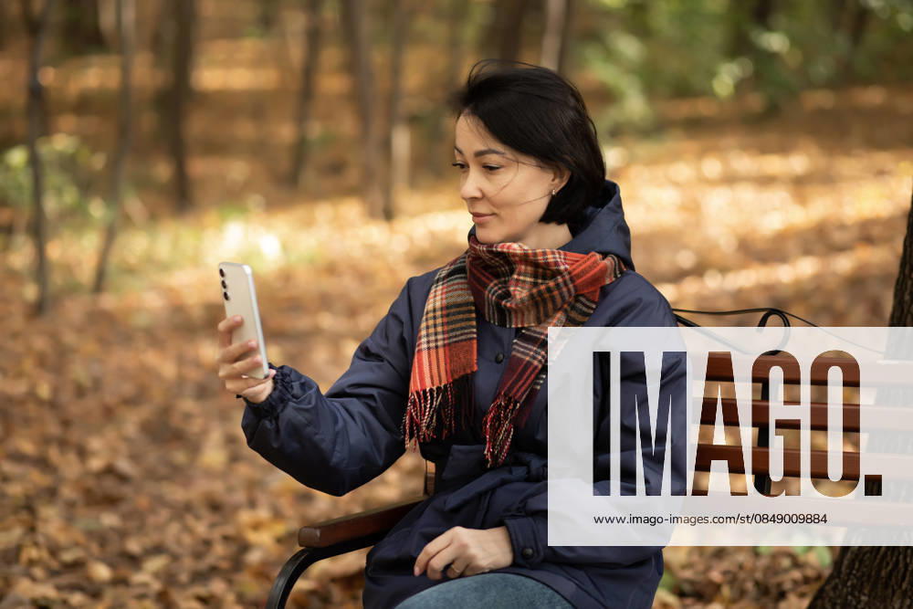 Woman enjoying autumn in park with smartphone Woman sitting on a park bench during autumn, holding a smartphone. Fallen leaves cover the ground, creating a warm, golden ambiance. The scene captures a peaceful, seasonal moment. Model Released ZulfiiaIshmukhametova_ID32598_1110457_006