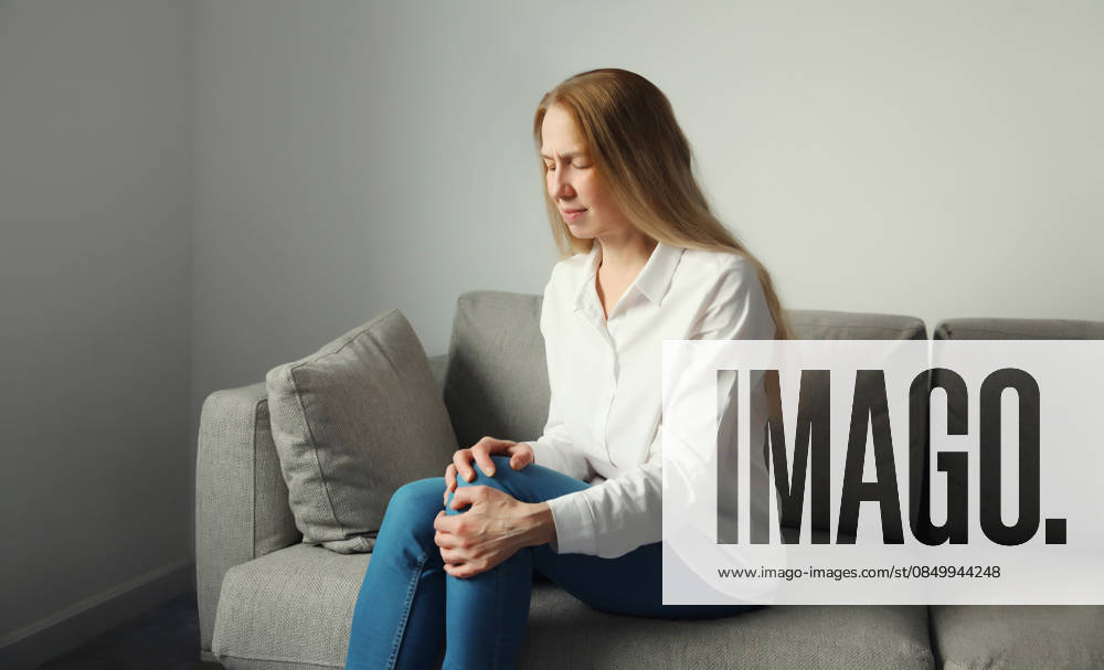 Frau leidet zu Hause unter Schmerzen im Knie und im Bein. Health care concept. Mature woman suffering from pain in knee rubbing leg sitting on couch at home. Old age, health problem and people concept (License=RF) ,model released, Symbolfoto