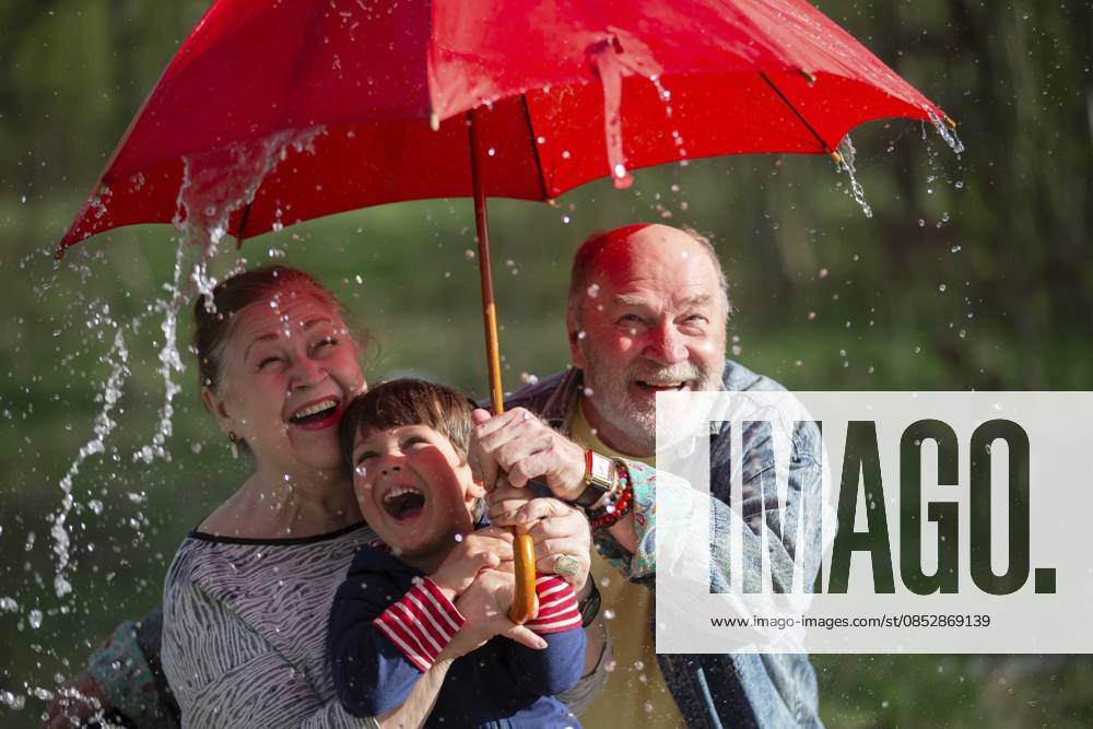 Großvater und Enkel halten einen Regenschirm in der Hand, und Großmutter schaut sie an. Die Szene ist lustig und unbeschwert. Familie im Regen Grandfather and grandson are holding an umbrella, and grandmother is looking at them. The scene is fun and light-hearted. family in the rain