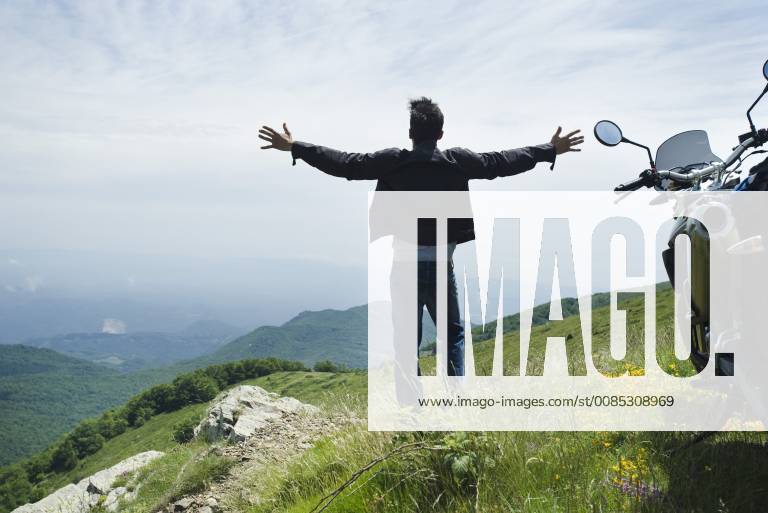 Man standing by motorcycle on top of mountain, arms outstretched