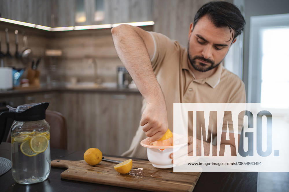 Ein Mann, der in der Küche Zitronen auspresst, um Entgiftungswasser oder Limonade zuzubereiten Mid adult man squeezing lemons in the kitchen preparing detox water or lemonade (License=RF) ,model released, Symbolfoto