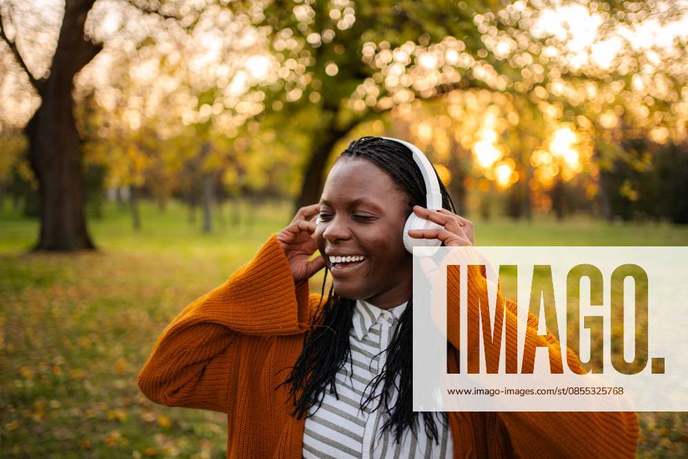 Eine fröhliche junge Frau, die im Herbst im Park Musik im Freien genießt Young black woman smiling, listening to music with headphones in an autumn park (License=RF) ,model released, Symbolfoto