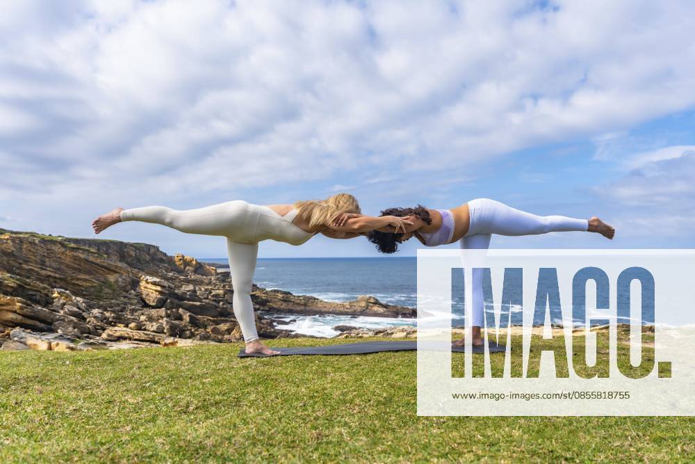 Zwei Frauen praktizieren im Freien auf einer grasbewachsenen Klippe mit Blick auf den Ozean Acro Yoga und halten zusammen eine tiefe Kriegerpose ein, die Balance, Teamwork und Wohlbefinden symbolisiert Two women practicing acro yoga outdoors on a grassy cliff overlooking the ocean, holding a deep warrior pose together, symbolizing balance, teamwork, and wellness