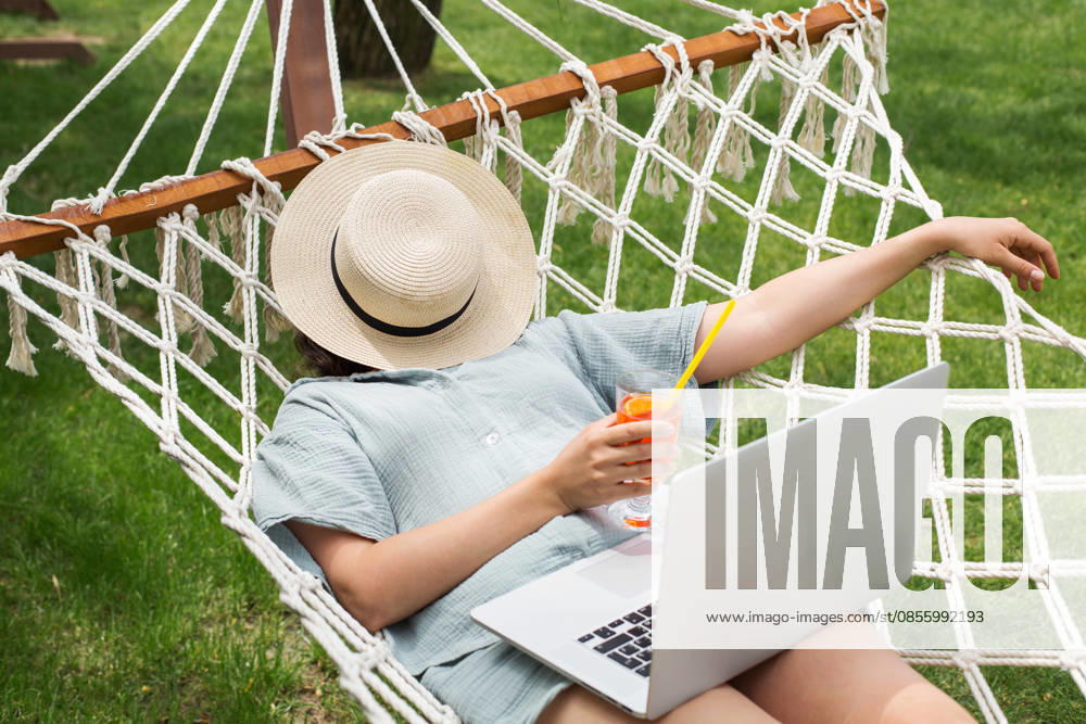 Frau, die sich mit ihrem Laptop in einer Hängematte entspannt und im Garten einen Sommercocktail in der Hand hält; Konzept für einen Urlaub mit Remote-Arbeit, das die Freiheit digitaler Nomaden, Freizeit und Wellness, mobile Produktivität sowie einen ausg Woman relaxing in hammock with laptop, holding summer cocktail in garden, remote work vacation concept expressing digital nomad freedom, leisure wellness, mobile productivity, balanced lifestyle (License=RF) ,model released, Symbolfoto