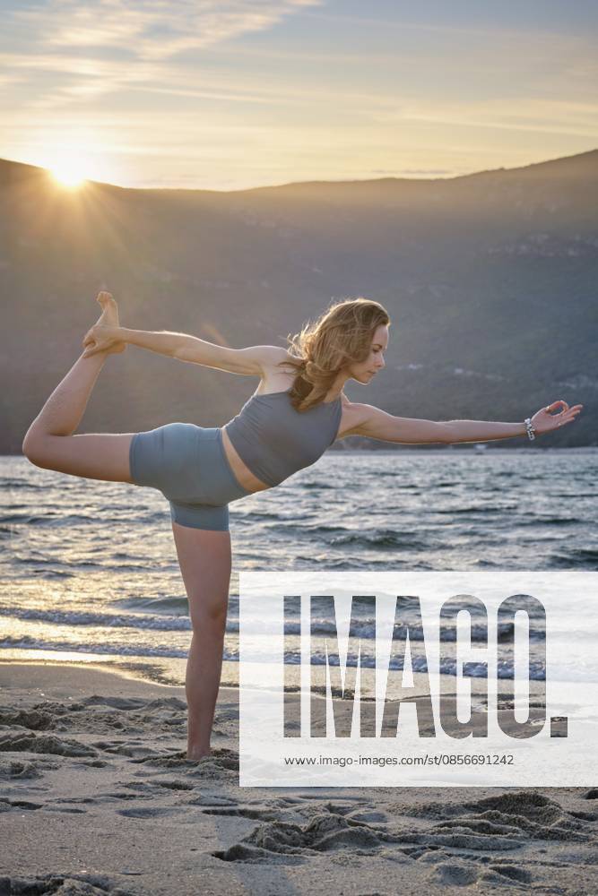 Eine Frau führt Natarajasana oder Dancer s Pose am Sandstrand bei Sonnenuntergang auf. Die ruhigen Meereswellen und die wunderschönen Berge bilden eine friedliche Kulisse für ihre Yoga Praxis A woman performs Natarajasana, or Dancer s Pose, on the sandy beach at sunset. The calm ocean waves and beautiful mountains create a peaceful backdrop for her yoga practice