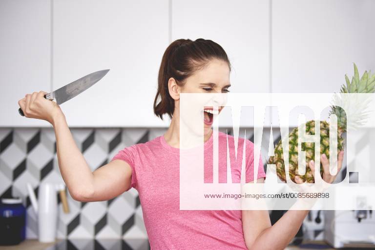 Portrait of screaming woman attacking pineapple with knife in the kitchen model released Symbolfoto property released ABIF00787