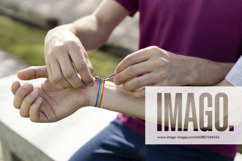 Nahaufnahme von zwei Männern, die ein Regenbogenbandarmband an ein Handgelenk binden, das LGBTQ+ Liebe, Stolz und Unterstützung symbolisiert Close-up of two men tying a rainbow ribbon bracelet on a wrist, symbolizing lgbtq+ love, pride, and support
